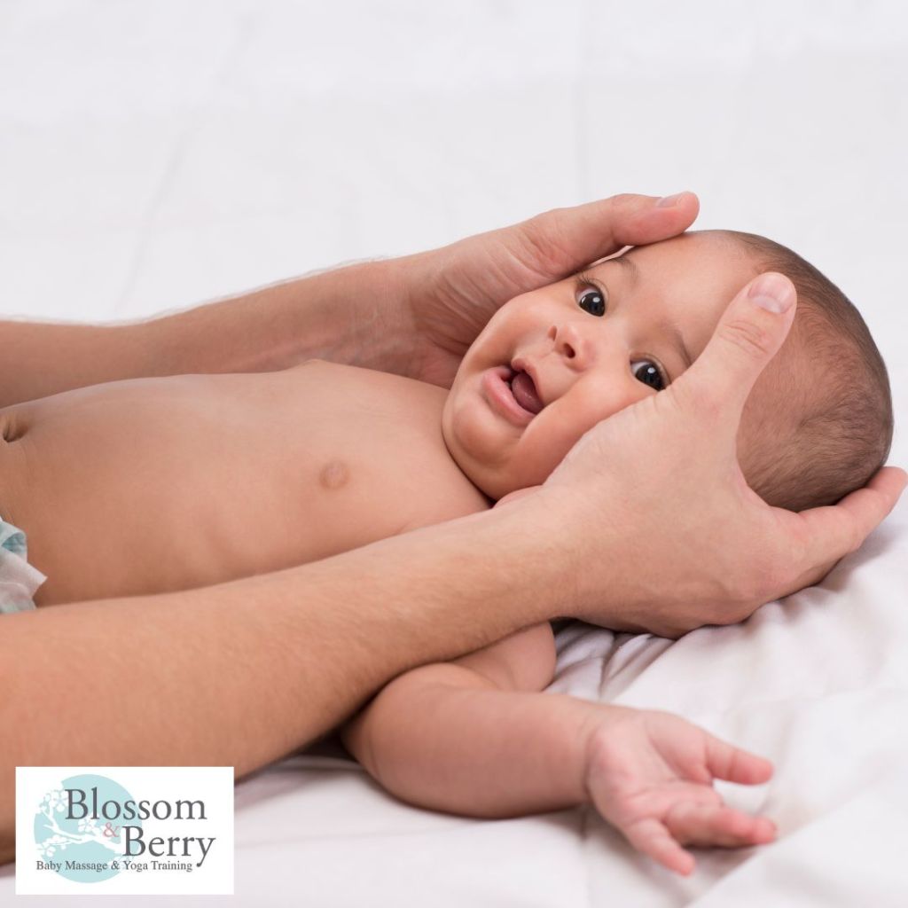 A parent gently framing a smiling baby's face with their hands while the infant lies comfortably on a soft white blanket.