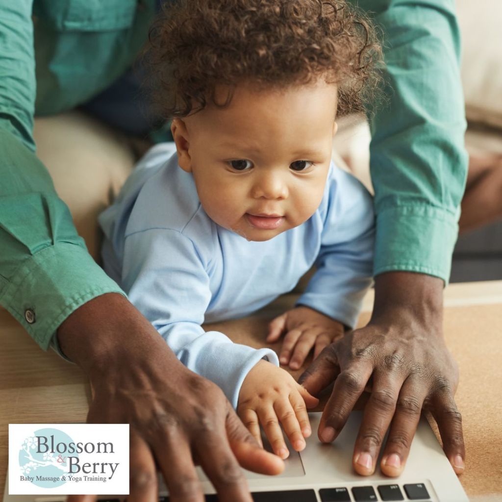Baby wearing a blue top leaning over a laptop keyboard with an adult guiding their hands